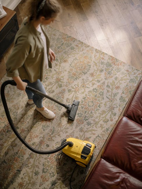 A young boy using a vacuum cleaner to clean a beige carpet in a living room while an older man is lying on a cream-colored sofa, appearing relaxed. The room features large windows with curtains allowi