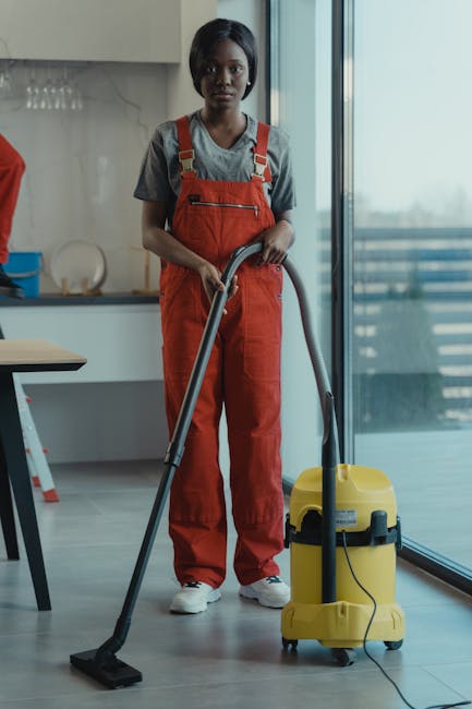 A female cleaner with light brown hair wearing a yellow uniform and a staff ID badge, smiling while holding a spray bottle in her right hand and a blue cleaning cloth in her left, standing in a well-l