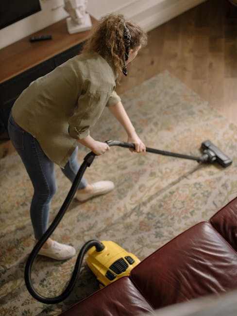 A person kneeling on a patterned area rug in a residential room, using a yellow portable vacuum cleaner with a black hose to perform surface cleaning and deep carpet cleaning. The individual is wearin