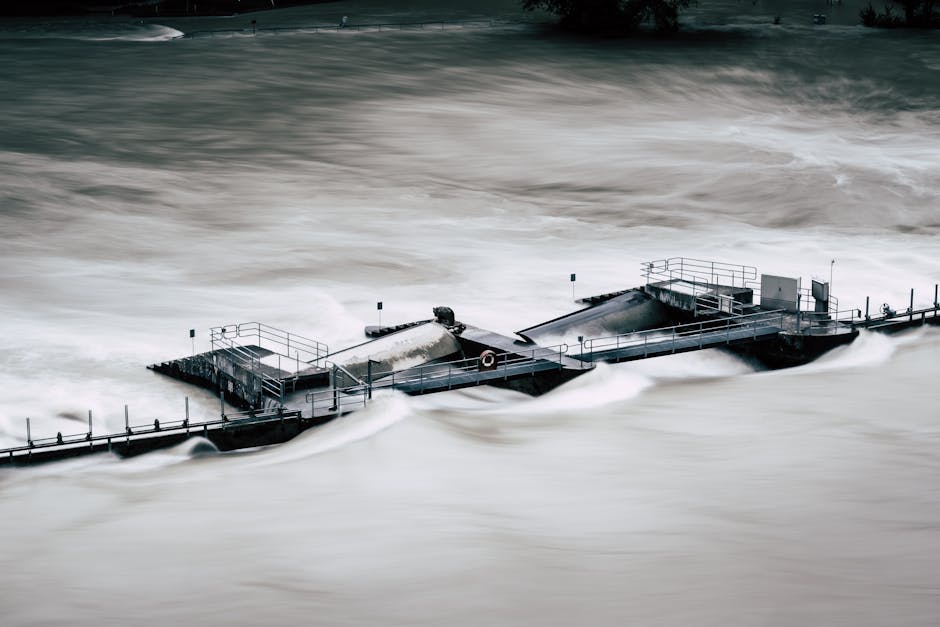 A flooded residential garden with water covering the grass and surrounding structures. The water reaches the base of a small house with a pitched roof, where part of a wooden fence and a white pole ar